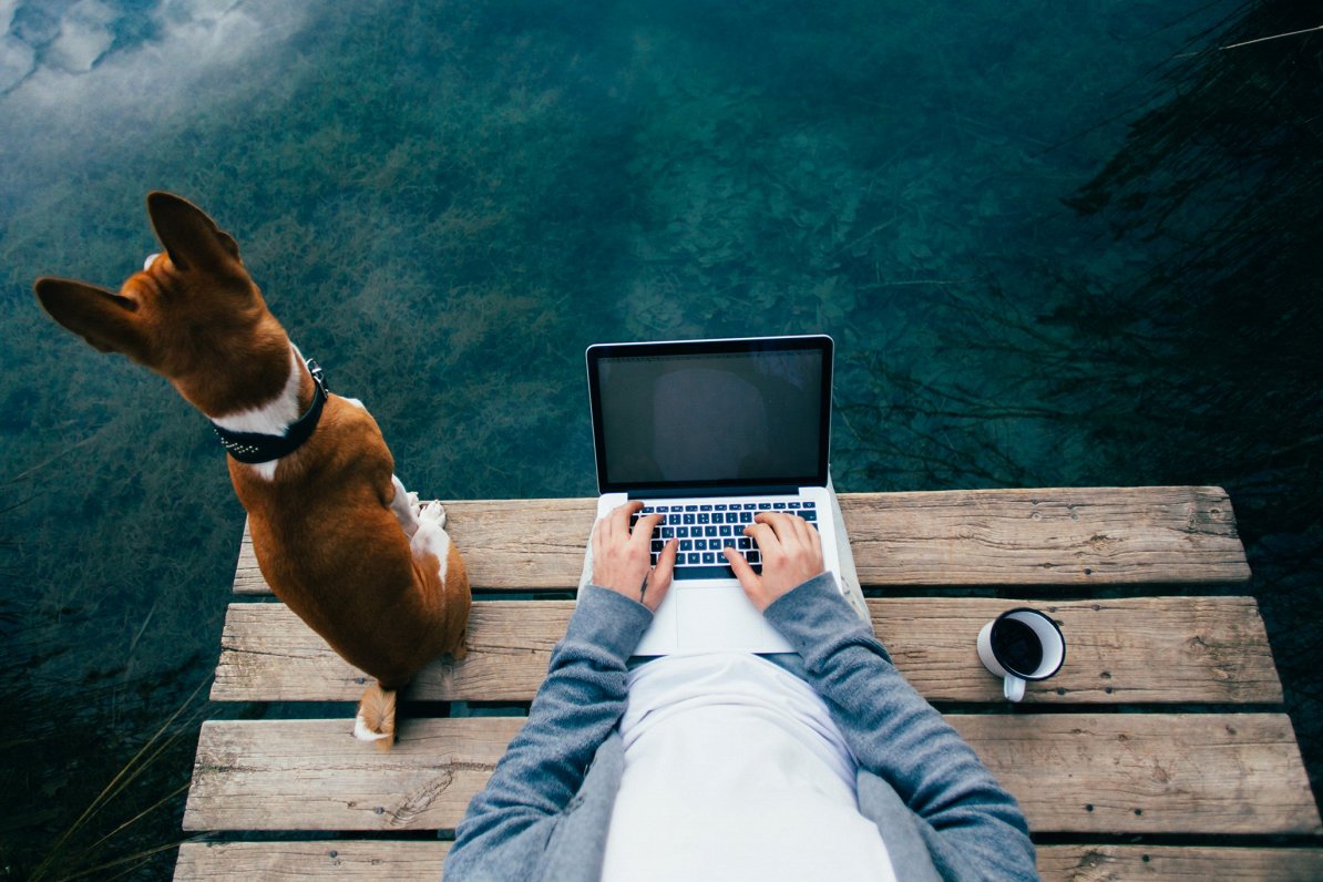 Datorlietotājs ar suni bauda rīta kafiju pie ezera