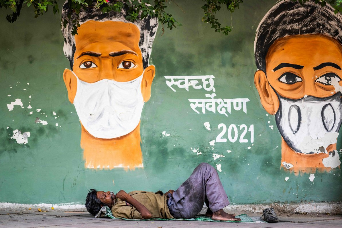 Indijā vīrietis atpūšas pie sienas gleznojuma, kas brīdina par Covid-19 briesmām
