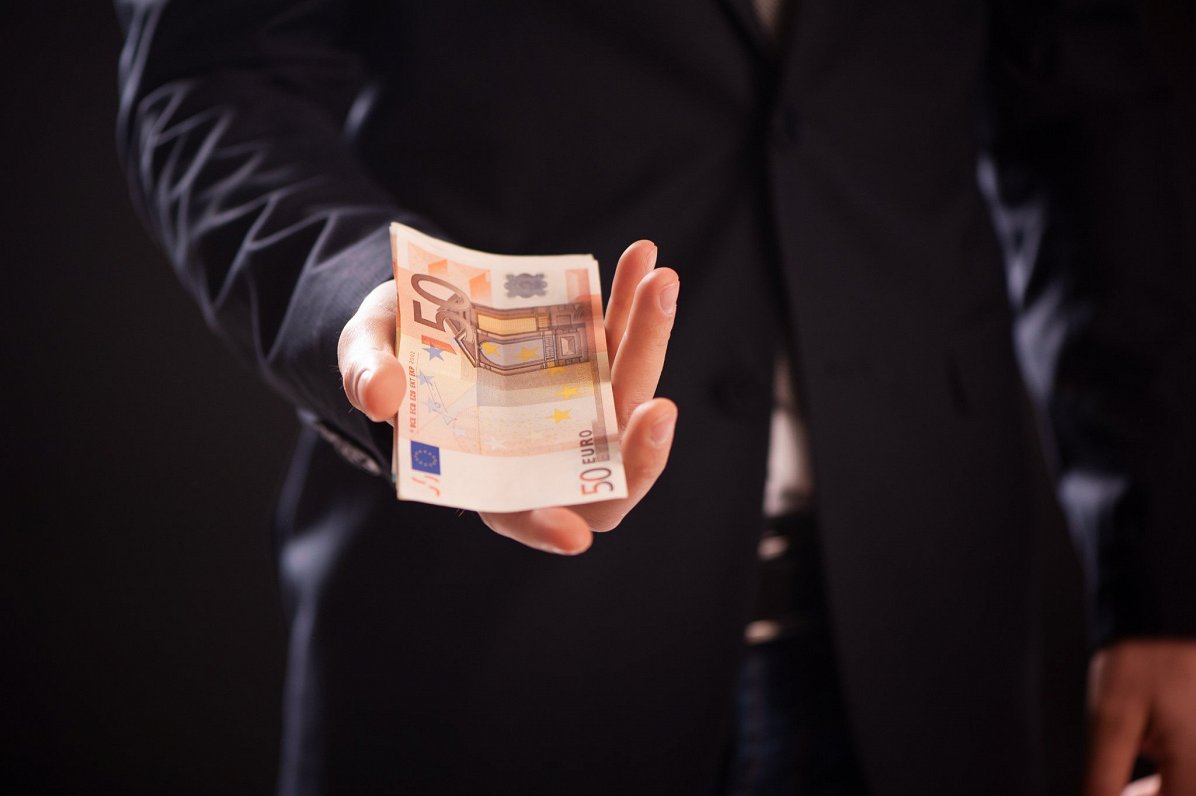Oficiāli ģērbts vīrietis ar eiro naudas banknotēm rokā. Attēls ilustratīvs.