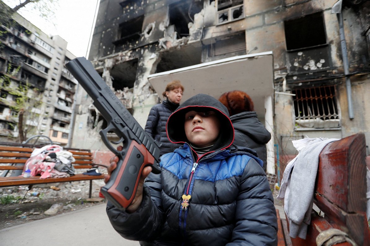 Bērns ar rotaļu pistoli pie sagrautas ēkas Mariupolē (21.04.2022)