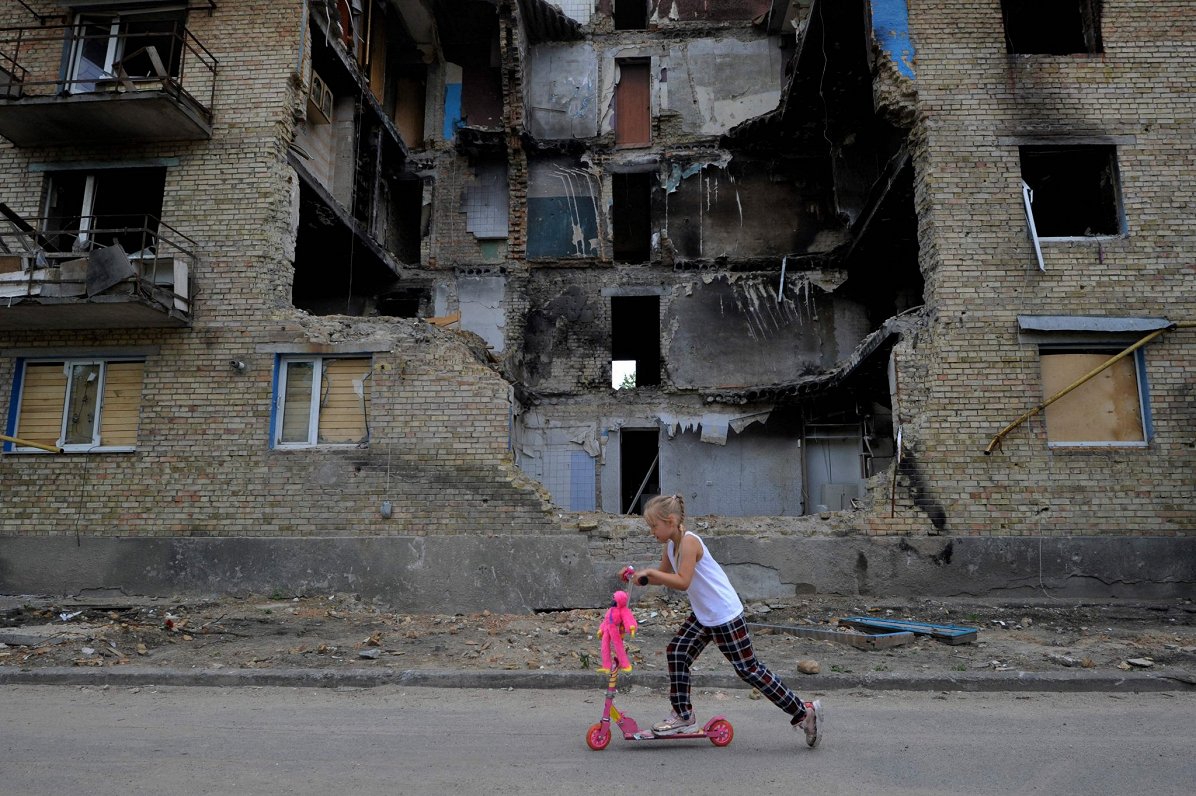 Ukrainā meitene ar skrejriteni brauc gar karadarbībā izpostītu māju