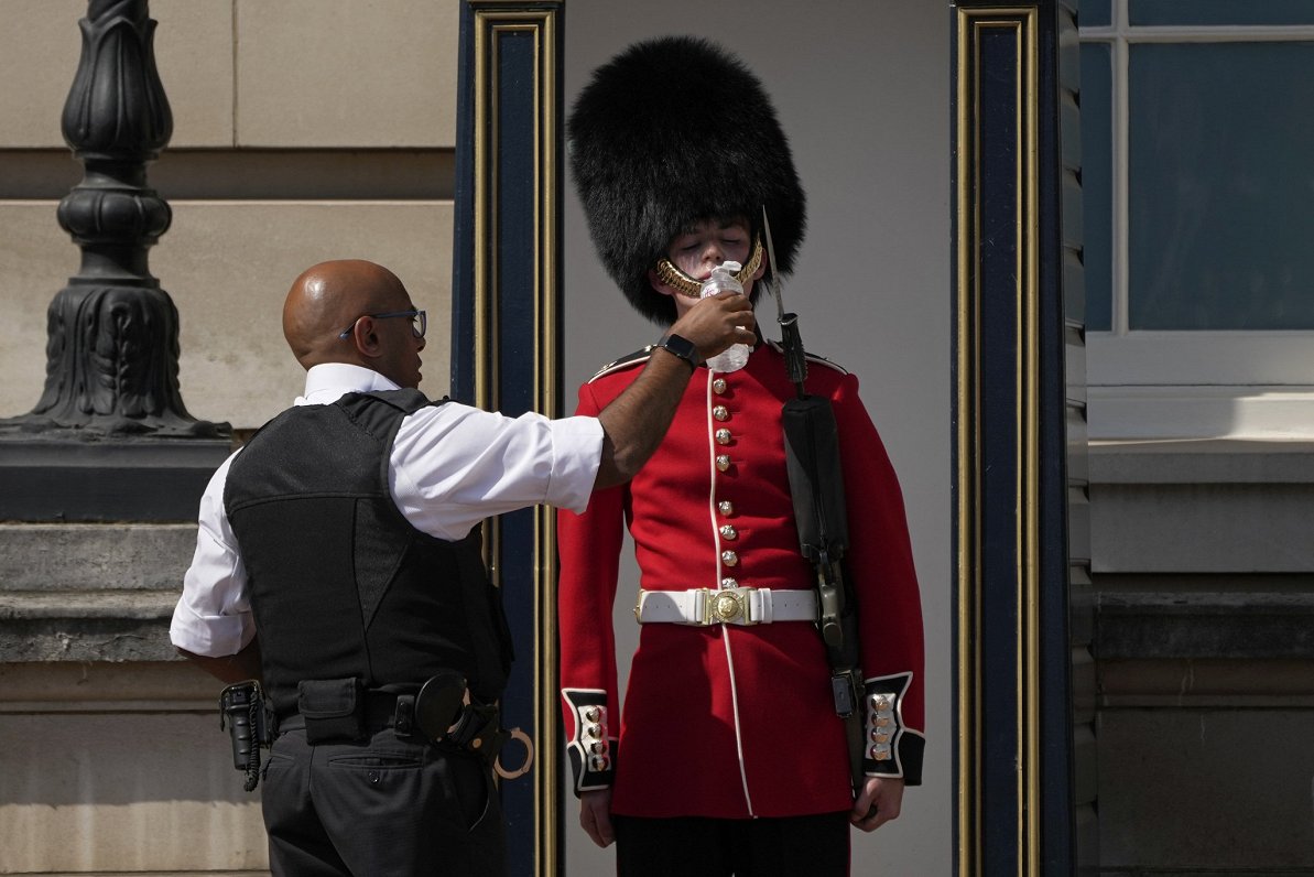 Britu policists pasniedz ūdeni sargam, kurš stāv godasardzē pie Bakingemas pils Londonā