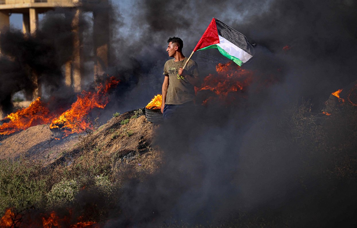 Palestīniešu jaunietis tur karogu pie degošām riepām protesta laikā pie robežas žoga ar Izraēlu uz a...