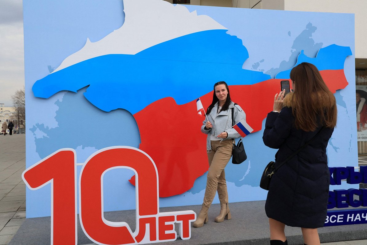 Krimas pilsētā Simferopolē sieviete pozē pie plakāta, kas cildina 10 gadu jubileju kopš Krimas iekļa...