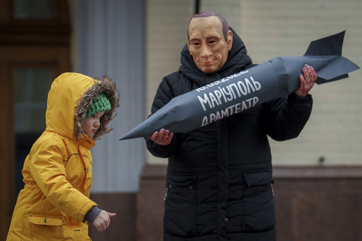 Krievijas vadoņa Vladimira Putina maskā tērpies vīrietis mītiņā Kijivā demonstrē raķetei līdzīgu vei...