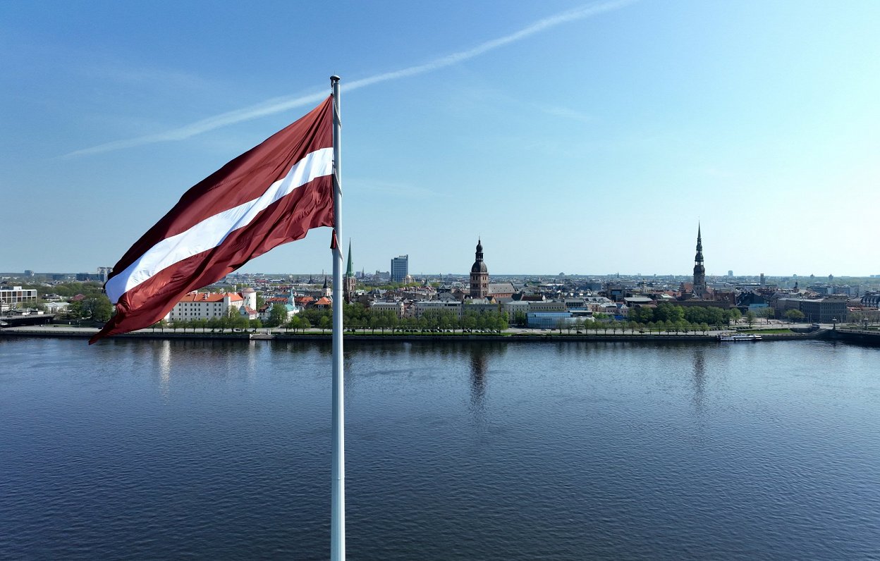 Latvijas karogs uz AB dambja un Rīgas panorāma.